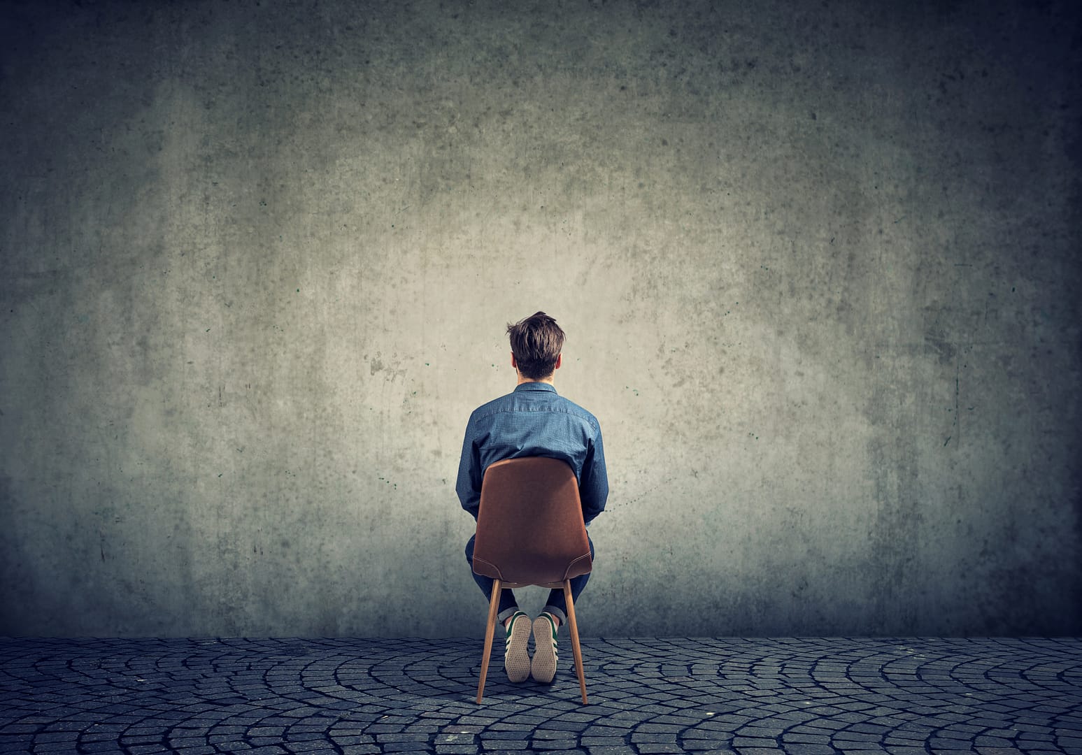 Eine Person sitzt rückwärts auf einem Stuhl, die Last der Depression ist spürbar, während sie auf eine kahle, strukturierte Wand auf einem Kopfsteinpflaster blickt.