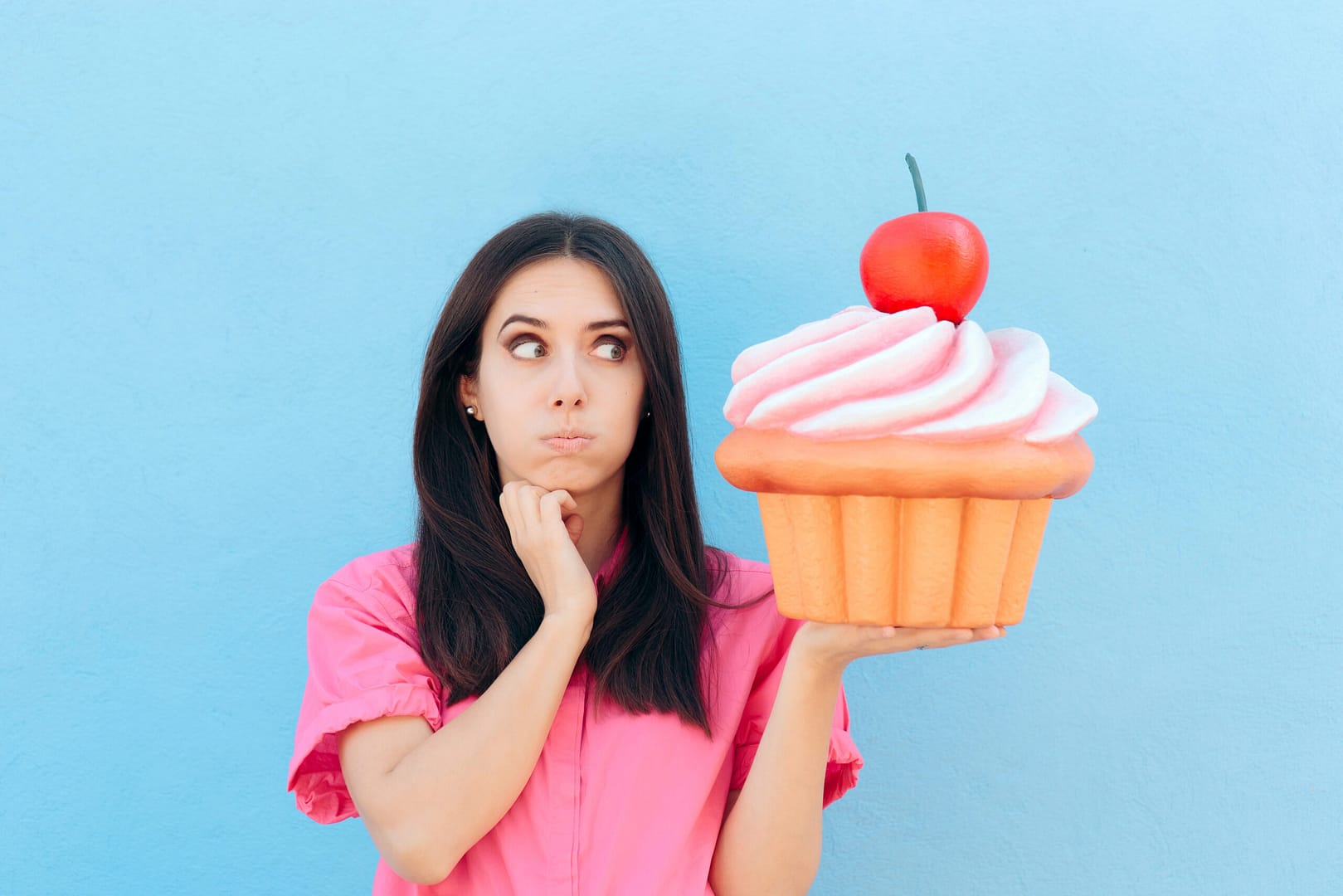 Eine Person in einem rosa Hemd hält einen großen falschen Cupcake mit einer Kirsche obendrauf und betrachtet ihn mit verwirrtem Gesichtsausdruck vor einem blauen Hintergrund.