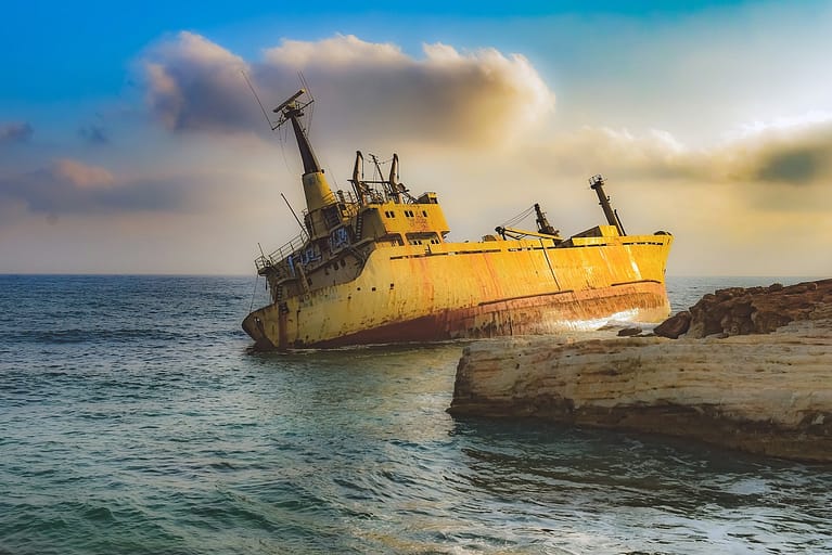 Ein nach rechts geneigtes, verrostetes Schiffswrack, das teilweise im Wasser in der Nähe einer felsigen Küste liegt, steht robust unter einem hellen Himmel mit vereinzelten Wolken – ganz ähnlich wie „Vertrauen in das deutsche Gesundheitssystem“.